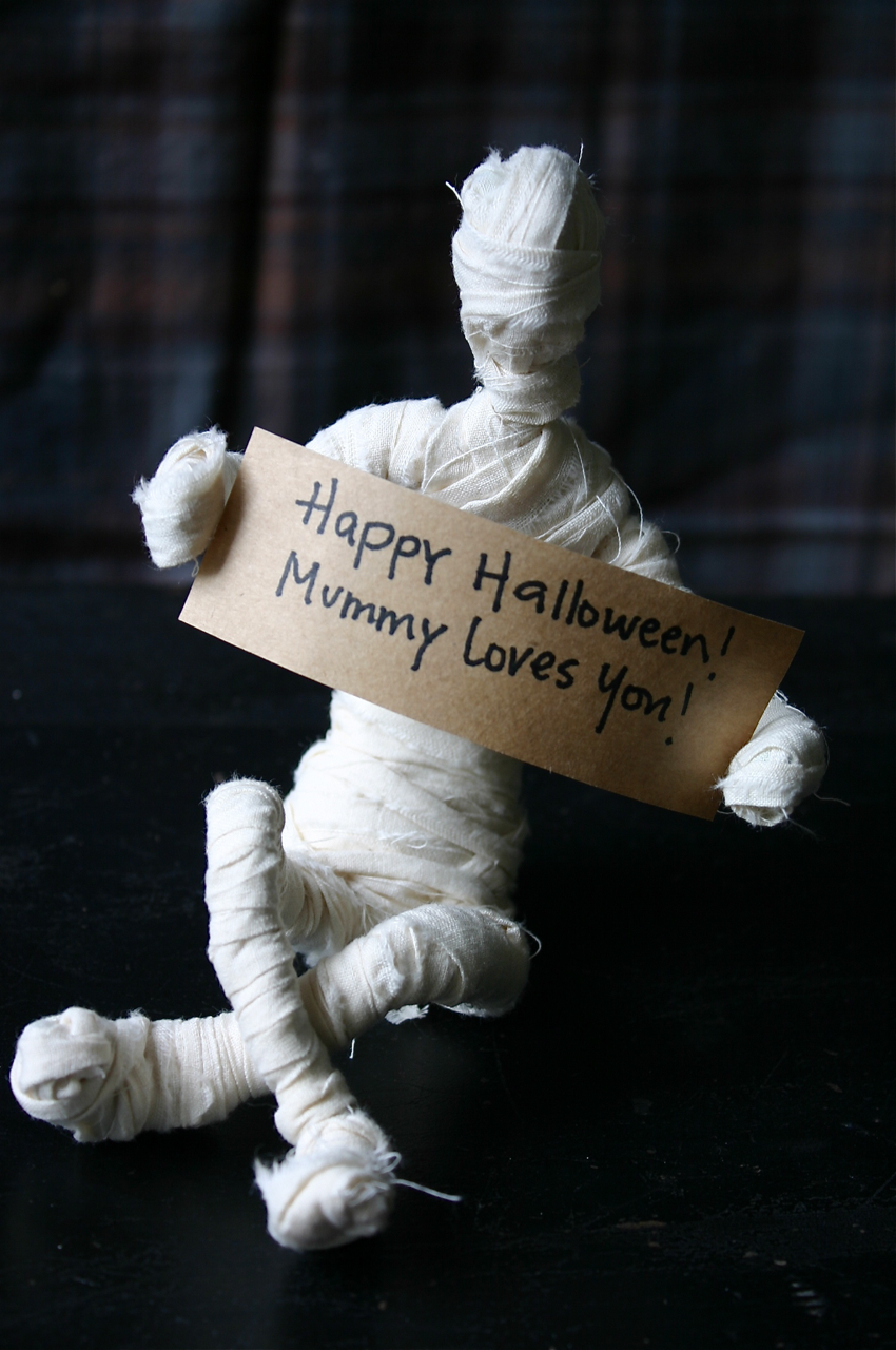Mummy in off white muslin holding up a white sign saying Happy Halloween and Mummy Loves You 