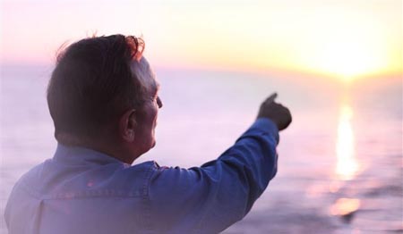 Back of a man pointing at the sun and he is near water. 