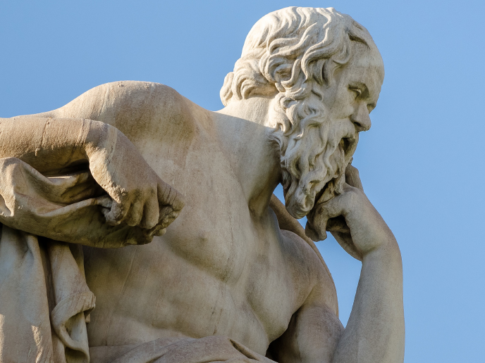 A stone statue of Socrates against a blue sky background. He is holding his head up with his hand in a thinking pose. 