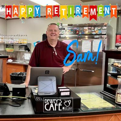 Cafe Manager Sam standing behind the Dewey's Counter with a "Happy Retirement Banner" hanging over his head