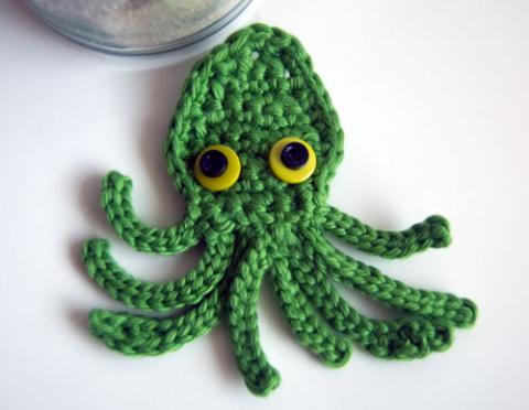 Green crochet squid/octopus with yellow and black eyes against a white background. 