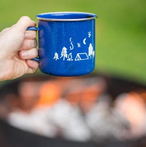 Person holding a blue enamel mug against a background with a campfire. 