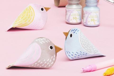Three paper roll birds in different colors against a pink background with two pots of paint also in the background with two paint pens on the lower right corner