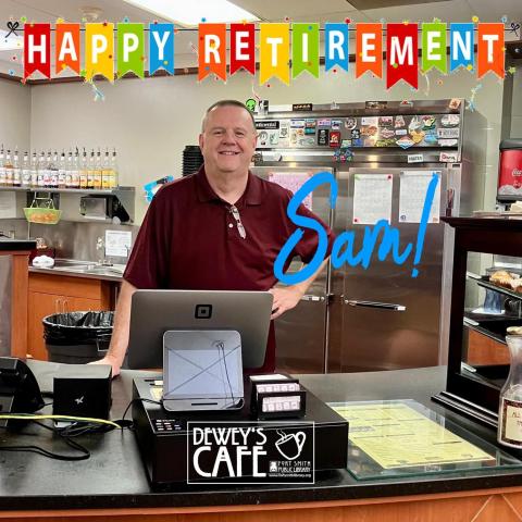 Cafe Manager Sam standing behind the Dewey's Counter with a "Happy Retirement Banner" hanging over his head