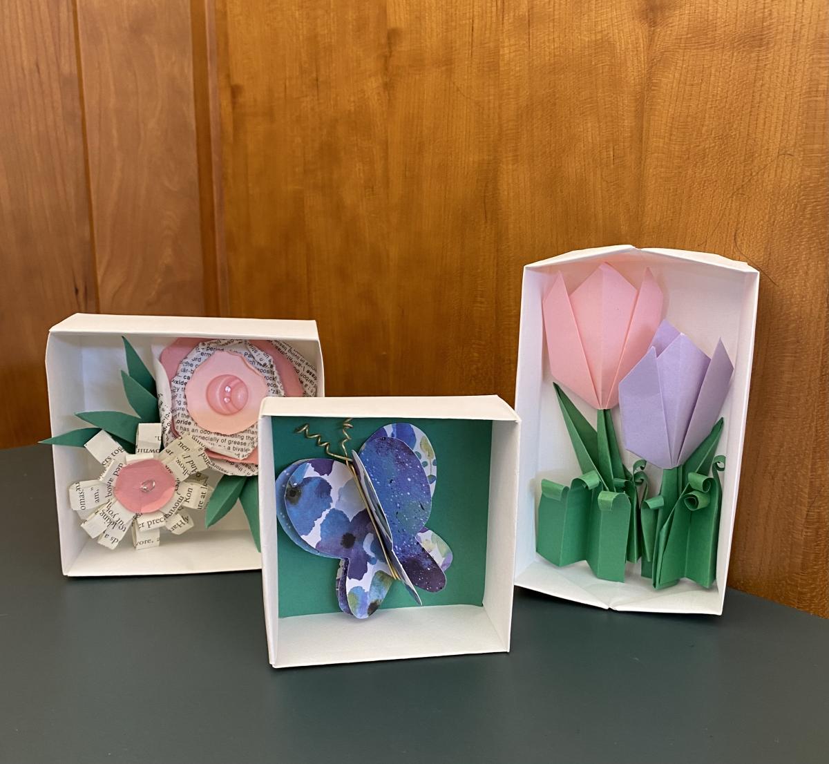 three white hand-crafted paper shadowboxes with paper flowers and butterflies inside them.